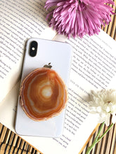 Load image into Gallery viewer, Natural agate phone grip presented on iPhone 8, laying on top of open book, framed by for and yellow flowers
