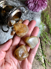 Load image into Gallery viewer, three large honey agate tumbled crystals lay in hand over boho background of fur, wood and flowers
