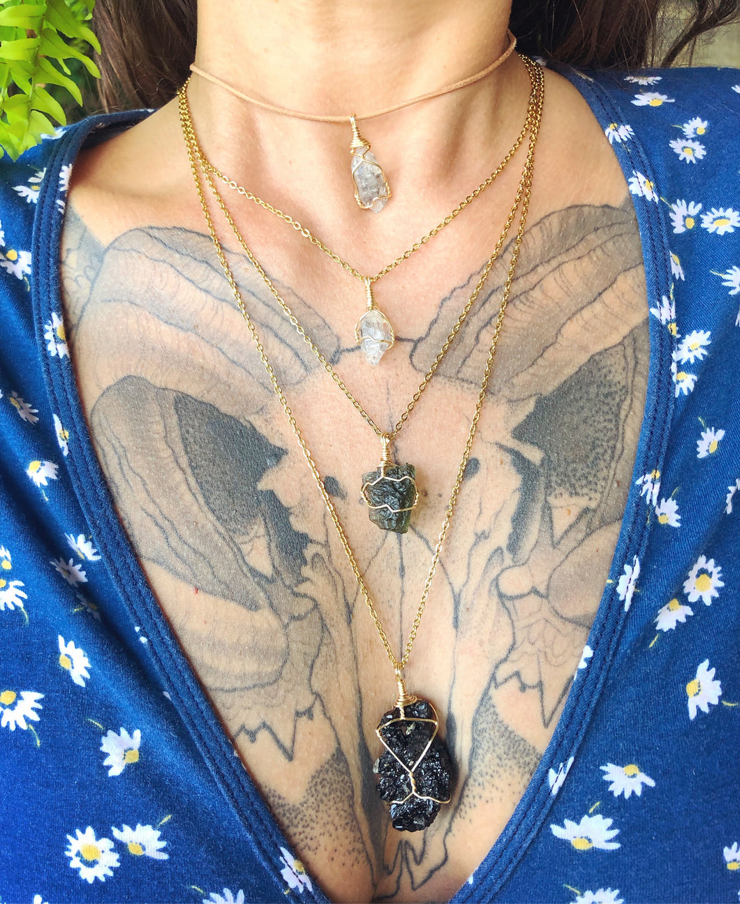 One herkimer diamond choker, one herkimer diamond necklace, one tourmaline necklace and one moldavite necklace all on gold chains layered over ram skull chest tattoo. Ferns hanging in the backgorund.