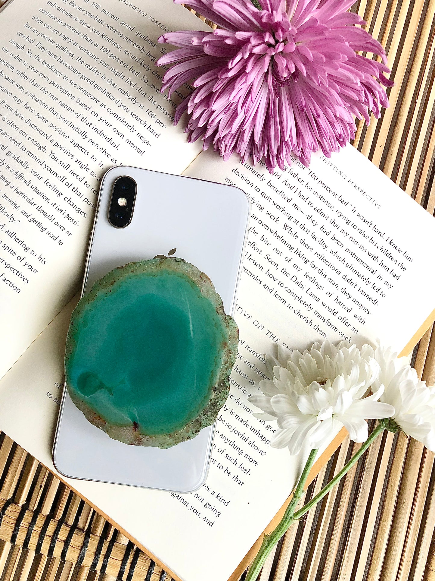 Natural greenish blue agate phone grip presented on iPhone 8, laying on top of open book, framed by for and yellow flowers