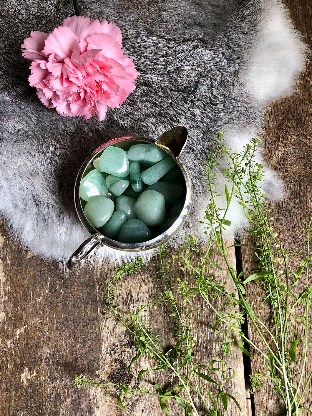 Aventurine tumbles sit in silver dish on top of fur and antique wood framed with greenery and pink flower