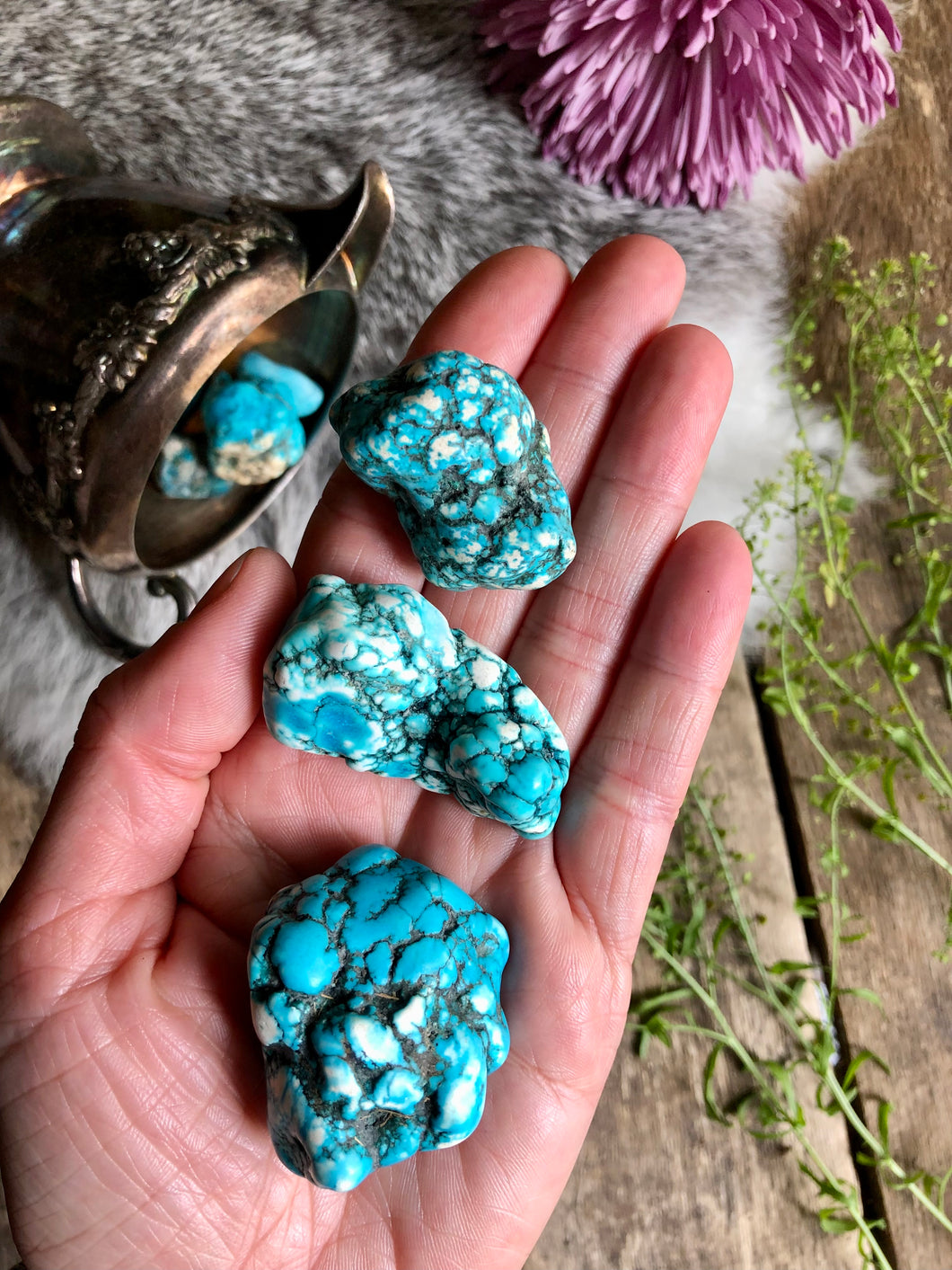 Large blue dyed howlite crystals lay in hand above boho background of fur, aged wood and pink and green foliage