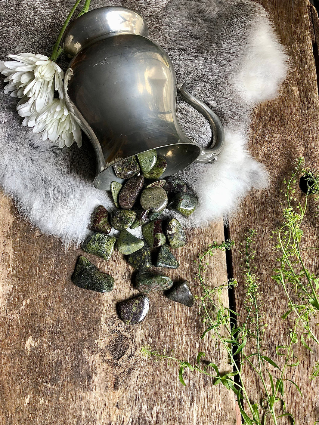 Dragon's Blood tumbled crystals spill out of silver antique gauntlet on top of fur and aged wood, framed with green and white foliage