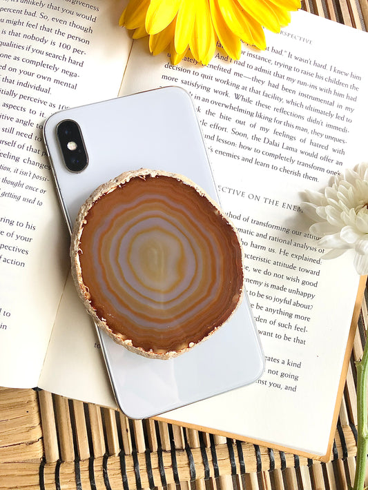 Natural brown agate phone grip presented on iPhone 8, laying on top of open book, framed by for and yellow flowers