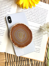 Load image into Gallery viewer, Natural brown agate phone grip presented on iPhone 8, laying on top of open book, framed by for and yellow flowers
