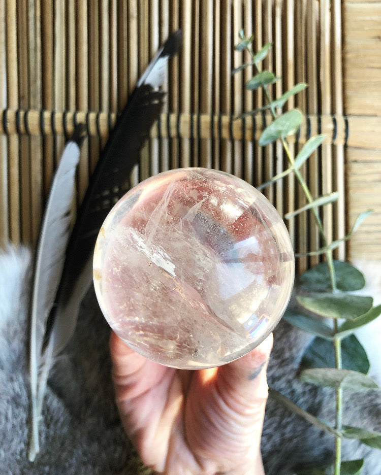 Clear quartz sphere in hand. Framed with feathers, fur and eucalyptus over wicker blurred in the distance.
